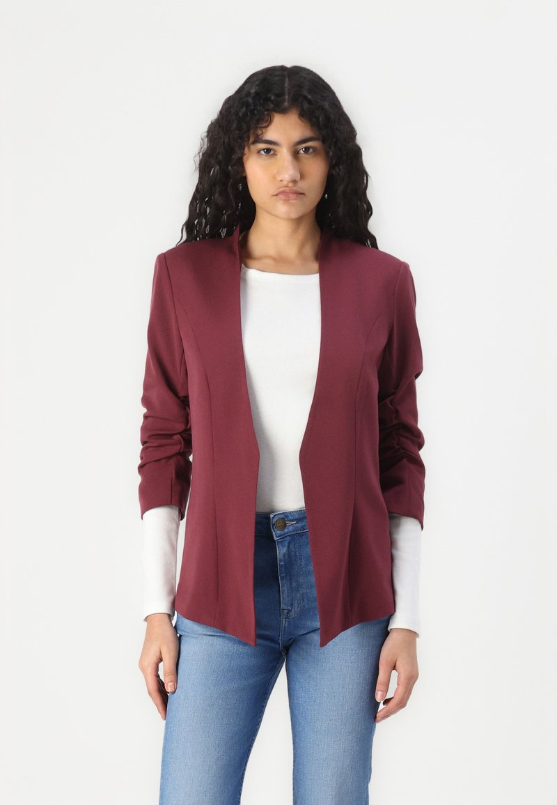 Burgundský blazer s otevřeným předním designem, dlouhými rukávy a vystuženými rameny, vrstvený přes bílou košili s dlouhým rukávem. Texturovaná tkanina.
