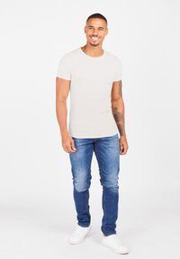 T-shirt aderente color beige chiaro, maniche corte; indossato con jeans slim-fit blu medio con macchie scolorite; abbinato a sneaker bianche.