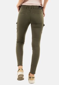 Pantalons cargo fins en vert olive, fabriqués à partir d'un matériau extensible. Présente des poches latérales et une taille haute. Portés avec des baskets beiges.
