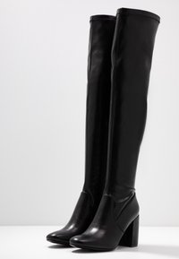 Botas de cuero negras por encima de la rodilla con una textura suave, punta redondeada, tacón alto y un detalle de cremallera en el lado interno.