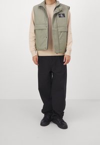 Olive zelená prošívaná vesta se zapínáním na patentky, dvěma předními kapsami a černou logo nášivkou, nošená přes béžový svetr a černé kalhoty s širokými nohavicemi. Černé tenisky.