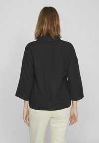 Črna, teksturirana srajca s kragnico in tridelnimi rokavi. Nosi se z svetlo obarvanimi hlačami, kar prikazuje sproščen silhuet.