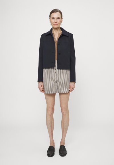 Veste navy courte avec trois boutons, chemise beige à boutons, shorts rayés et mocassins noirs. Tissu lisse avec un design structuré.