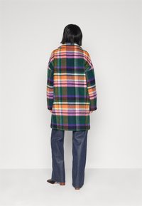 Person står med ryggen til iført en flerfarvet ternet frakke, blå jeans og sorte spidse sko mod en simpel hvid baggrund.