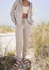 Beżowa bluza z kapturem, dopasowane dresy z elastycznymi mankietami oraz jasnobrązowe sandały z dwoma paskami. Gładka bawełniana tekstura, luźny krój.