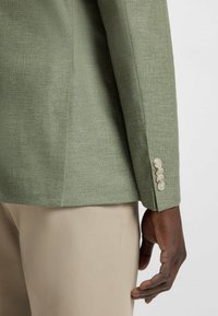 Lichtgroene blazer met een gestucte afwerking en knopen bij de manchetten, gecombineerd met beige broeken. Focus op op maat gemaakte ontwerpelementen.