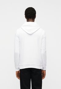 Άνδρας που στέκεται γυρίζοντας πλάτη, φορώντας μονόχρωμο λευκό φούτερ με κουκούλα και μαύρο παντελόνι, με φόντο λευκό και μονόχρωμο.