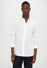 Camisa branca de manga longa, com botões, feita de tecido leve e com colarinho clássico. Apresenta punhos arregaçados e uma bainha reta.