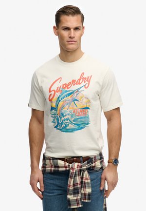 Homme portant un T-shirt blanc avec un poisson marlin coloré et l'inscription « Superdry Atlantic Coast », une chemise à carreaux attachée autour de la taille, un jean bleu et une montre-bracelet.