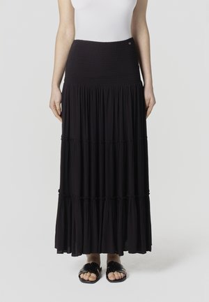 Naine kannab kõrge vöökohaga musta kihilist maksiseelikut, valget topi ja musti liugjalanõusid, seistes tavalise tausta ees.