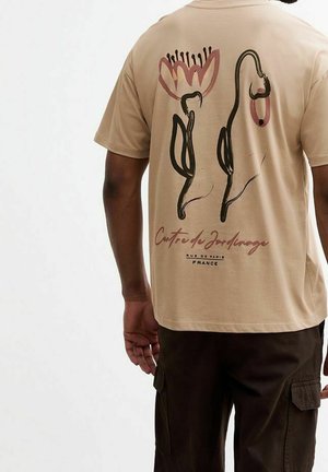 T-shirt en coton couleur fauve avec un motif graphique de fleur et formes abstraites au dos, avec le texte "Centre de Jardinage".