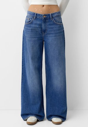 Jean bleu à jambes larges porté avec des baskets blanches et un top court blanc, visible de la taille aux pieds sur un fond uni.