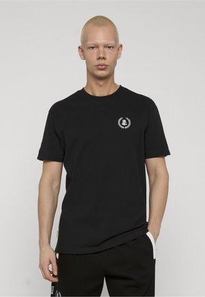 Ung person med kort blondt hår iført en svart t-skjorte og svarte bukser, stående foran en ensfarget lys bakgrunn.