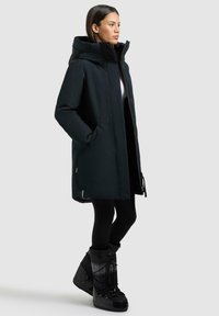 Schwarze isolierte Parka mit Kapuze, seitlichen Taschen und geradem Schnitt. Kombiniert mit schwarzen Schneestiefeln im Schnürdesign.