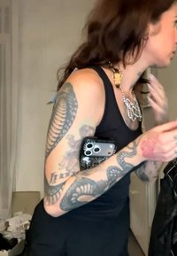 Personne avec des tatouages de serpent et de crâne sur les bras, portant un haut noir sans manches et plusieurs colliers, tenant un téléphone contre son côté.