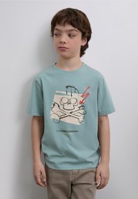 T-shirt en coton bleu clair arborant un motif de tête de mort et os croisés avec des accents rouges. Manches courtes et coupe décontractée, associé à un pantalon beige.