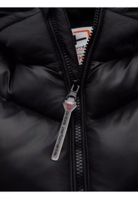 Dettaglio di un piumino nero con chiusura a zip, texture lucida, materiale leggero e un'etichetta con logo e hardware in argento.