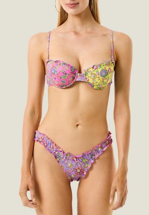 Donna che indossa un bikini floreale multicolore con bordi smerlati e spalline regolabili, in piedi davanti a uno sfondo chiaro uniforme.