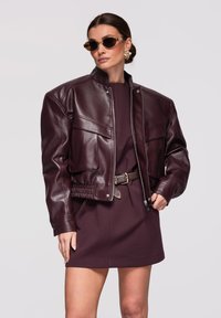 Femme portant une veste en cuir brun foncé surdimensionnée, une robe assortie avec ceinture, de grandes boucles d'oreilles en or et des lunettes de soleil, posant devant un fond uni.