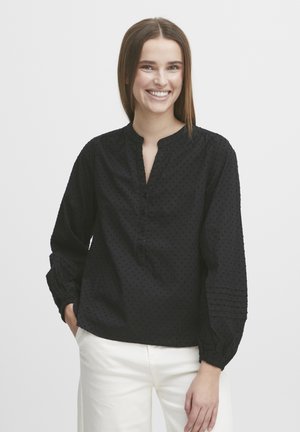 Black long-sleeve blouse with a v-neck, textured polka dot pattern, front button closure, and gathered cuffs. Paired with light-colored pants.