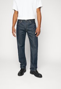 Mörkblå denimjeans med rak ben, fem fickor och kontrasterande sömnadsdetaljer, tillsammans med en vit t-shirt och svarta skor.