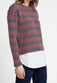 Haut à manches longues en maille avec un motif texturé en rouge, marine et blanc. Look superposé avec un ourlet blanc et un col arrondi.