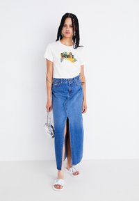 T-shirt blanc avec un graphisme coloré "Levi's", jupe en denim bleu taille haute avec une fente à l'avant, sac en chaîne argentée et sandales blanches à enfiler.