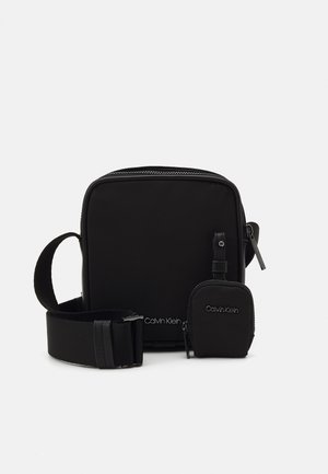 Zwarte compacte crossbodytas met verstelbare band en klein aangehecht zakje, beide bedrukt met Calvin Klein-logo's.