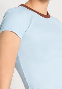 Světle modré žebrované tričko s hnědým kontrastním kulatým výstřihem. Krátké rukávy s přiléhavým střihem a hladkou texturou.