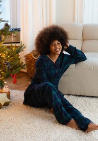 Groene en marineblauwe geruite pyjama met rode accenten, zachte stof, lange broek, zittend op een tapijt bij een kerstboom en bank.