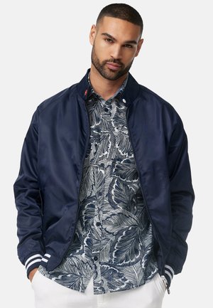 Chaqueta bomber azul marino con puños de rayas blancas, textura suave, cierre de cremallera, usada sobre una camisa de manga corta con un patrón en azul y blanco.
