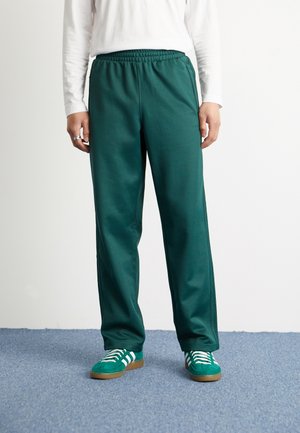 Pantaloni sportivi verdi in tessuto liscio, dotati di vita elasticizzata e tasche laterali. Abbinati a scarpe verdi con lacci bianchi.