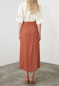 Gonna midi color ruggine con pois bianchi, realizzata in tessuto morbido. Presenta un design a vita alta e una silhouette fluida, abbinata a sandali color pesca.