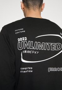 Schwarzes Langarmshirt mit weißem Text, darunter "2022 UNLIMITED" und verschiedenen grafischen Elementen, mit rundem Halsausschnitt und weicher Textur.