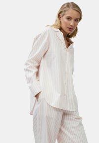 Frau in einem weißen Pyjama-oberteil mit Knöpfen und einer Hose mit dünnen rosa vertikalen Streifen, die die Hände in den Taschen hat und in die Kamera schaut.