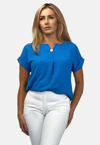 Niebieska bluzka z krótkim rękawem i dekoltem w serek, z detalem złotego guzika, połączona z białymi dopasowanymi spodniami. Tekstura tkaniny z podwiniętymi rękawami.