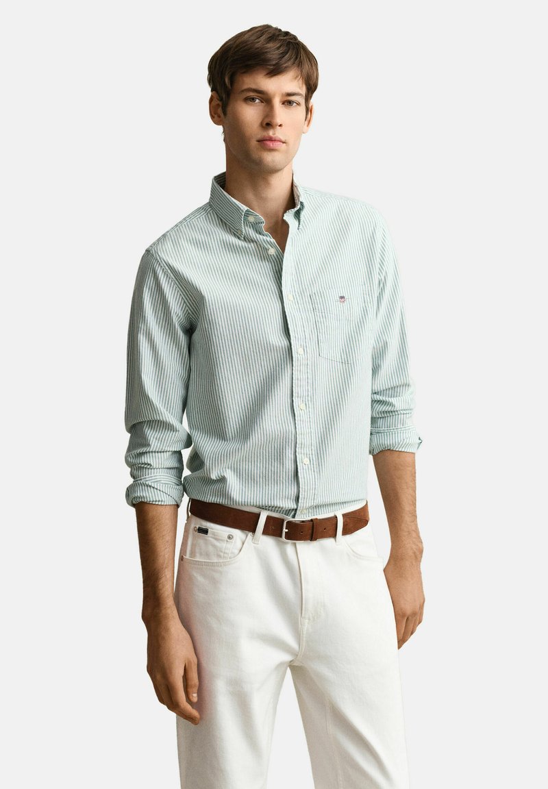 Giovane uomo che indossa una camicia a righe verde chiaro con maniche arrotolate, pantaloni bianchi e cintura marrone, in piedi davanti a uno sfondo semplice.