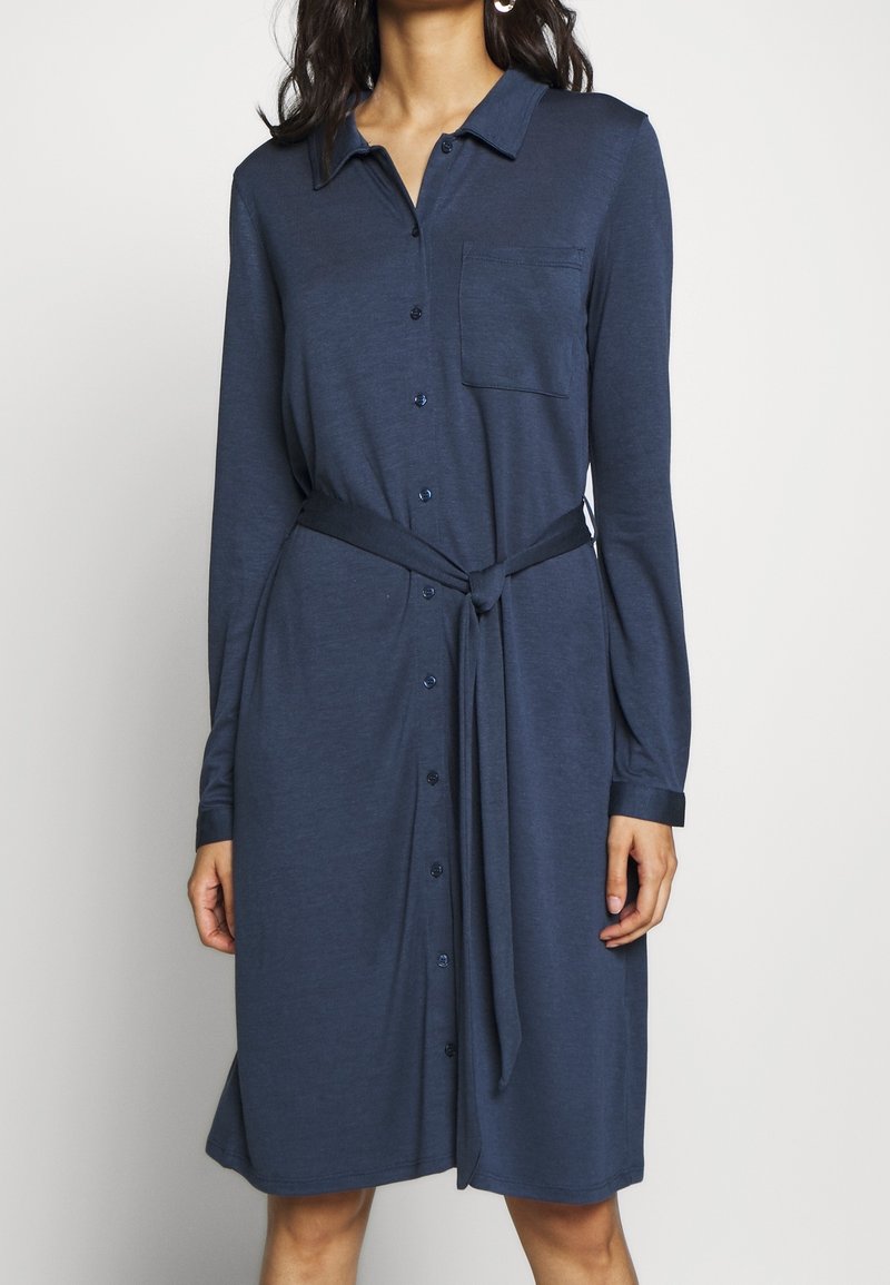 Robe boutonnée bleu marine avec manches longues, poche poitrine et ceinture nouée à la taille. Tissu doux avec une texture lisse et une coupe décontractée.