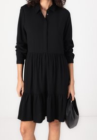 Abito camicia nero con maniche lunghe, chiusura frontale con bottoni, orlo a volant a strati e tessuto morbido. Tenuto con una borsa nera.