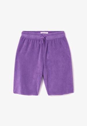 Dijete s kovrčavom kosom stoji, nosi pretjerano veliku narančastu majicu, ljubičaste kratke hlače i svijetlosmeđe cipele bez vezica na jednostavnoj pozadini.