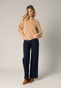 Beige fuzzy trui met wijde mouwen, gecombineerd met donkerblauwe wijde jeans en tan enkellaarsjes. Neutrale achtergrond benadrukt de outfit.
