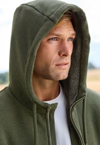 Olivgrüner Kapuzenpullover mit Reißverschluss, weicher Textur und fleecegefüttertem Innenfutter. Glattes Gewebe mit Kontrastnähten.