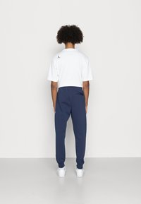 Persona in piedi di fronte a una parete bianca, vestita con una maglietta bianca con un piccolo logo sulla parte superiore della schiena e pantaloni jogger blu navy con scarpe da ginnastica bianche.