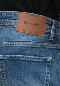Οι μπλε τζιν παντελόνες διαθέτουν ένα καφέ δερμάτινο πάτσα με την ένδειξη "ONLY & SONS" στο πίσω μέρος, με ραφές και μια ελαφρώς φθαρμένη υφή.
