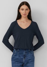 Marineblaues Langarm-V-Ausschnitt-Shirt aus Baumwollmischung, lockere Passform, mit einer glatten Textur und subtilen Nähdetails. Kombiniert mit hoch taillierten Jeans.