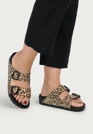 Feet wearing black cropped pants and leopard-print slip-on sandals with two black buckle straps.