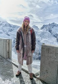 Žena v vzorovaném dlouhém kabátu, s růžovou čepicí a turistickými botami stojí na zasněženém horském výhledu s zasněženými vrcholky v pozadí.