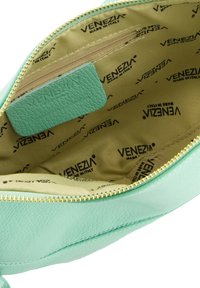 Mintgrøn lædertaske indvendigt med lynlåslomme, foret med beige stof, der viser det gentagne "VENEZIA" logo-mønster. Guld lynlåsdetalje.