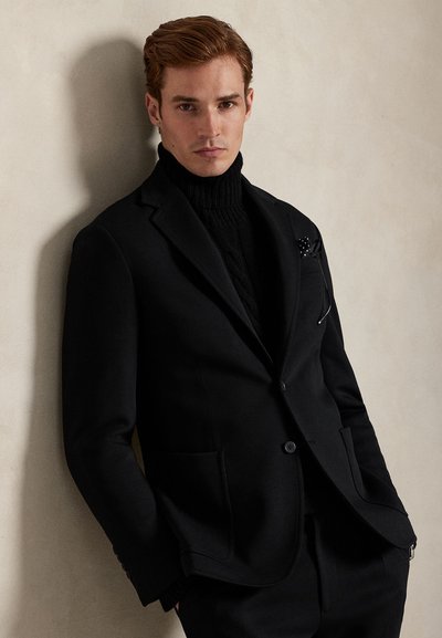 Μαύρο σακάκι προσαρμοσμένης γραμμής με τσέπες, φορεμένο πάνω από μαύρο πλεκτό ζιβάγκο. Το μοντέλο έχει κοντά μαλλιά και σοβαρή έκφραση, σε ουδέτερο φόντο.