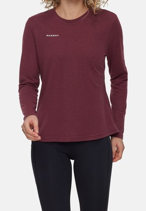 Vrouw die een lang mouw bordeaux top draagt met het "Mammut"-logo op de borst, gecombineerd met zwarte strakke broek, staand voor een effen achtergrond.